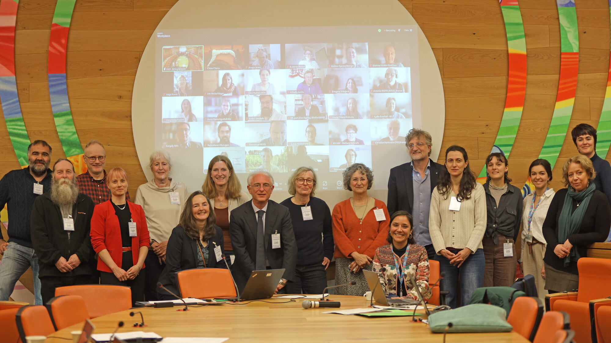 Group picture of members at the 12th Plenary Meeting 2025 at FAO headquarters in Rome. 18 people are standing in front of a big screen with an ongoing online-meeting with many more online participants..